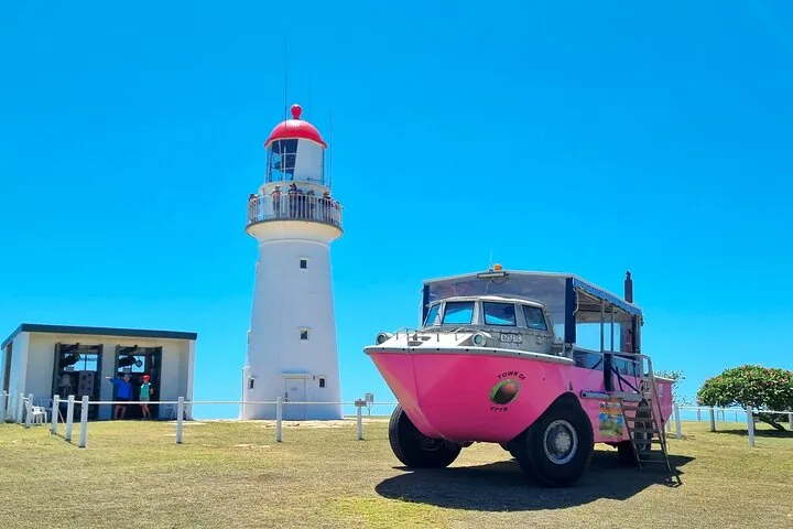 Ein rosa Amphibienfahrzeug, geparkt neben einem weißen Leuchtturm unter blauem Himmel